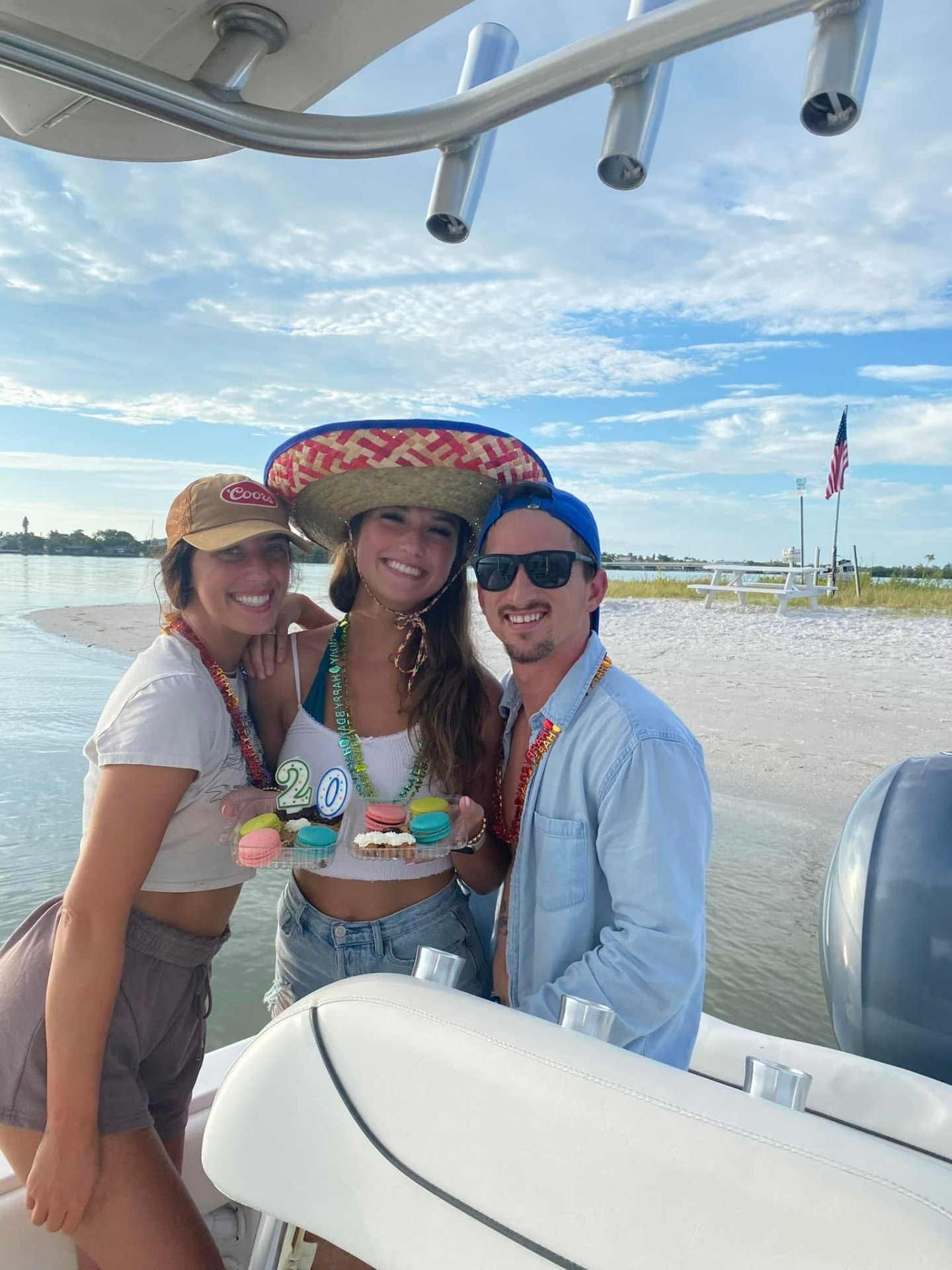 Annalyse, her sister in a sombrero, and Leon holding macarons with a 20 candle on a boat by a sandbar
