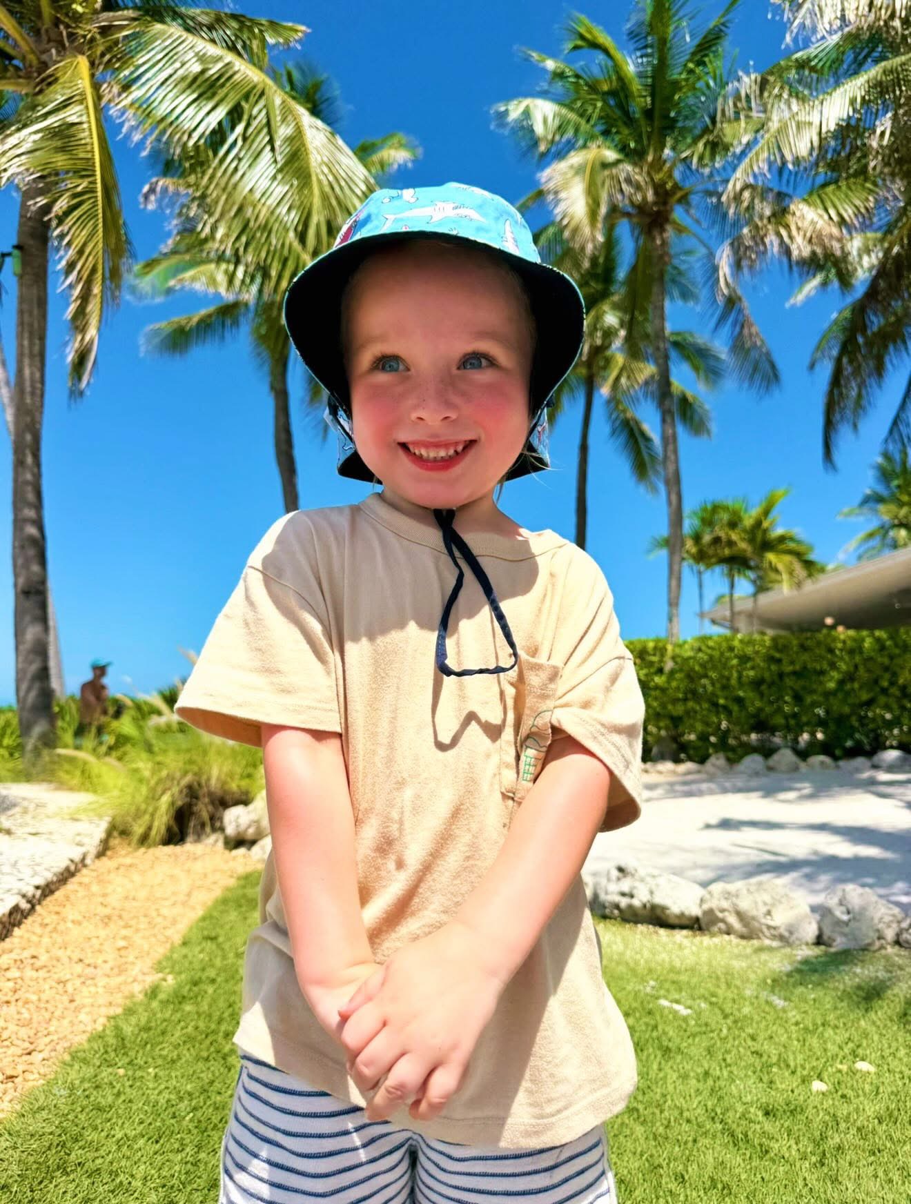 A smiling toddler in a shark bucket hat standing under palm trees
