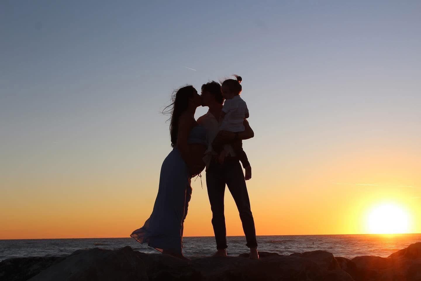 Silhouette of Annalyse and Leon kissing while holding their toddler on rocks at sunset