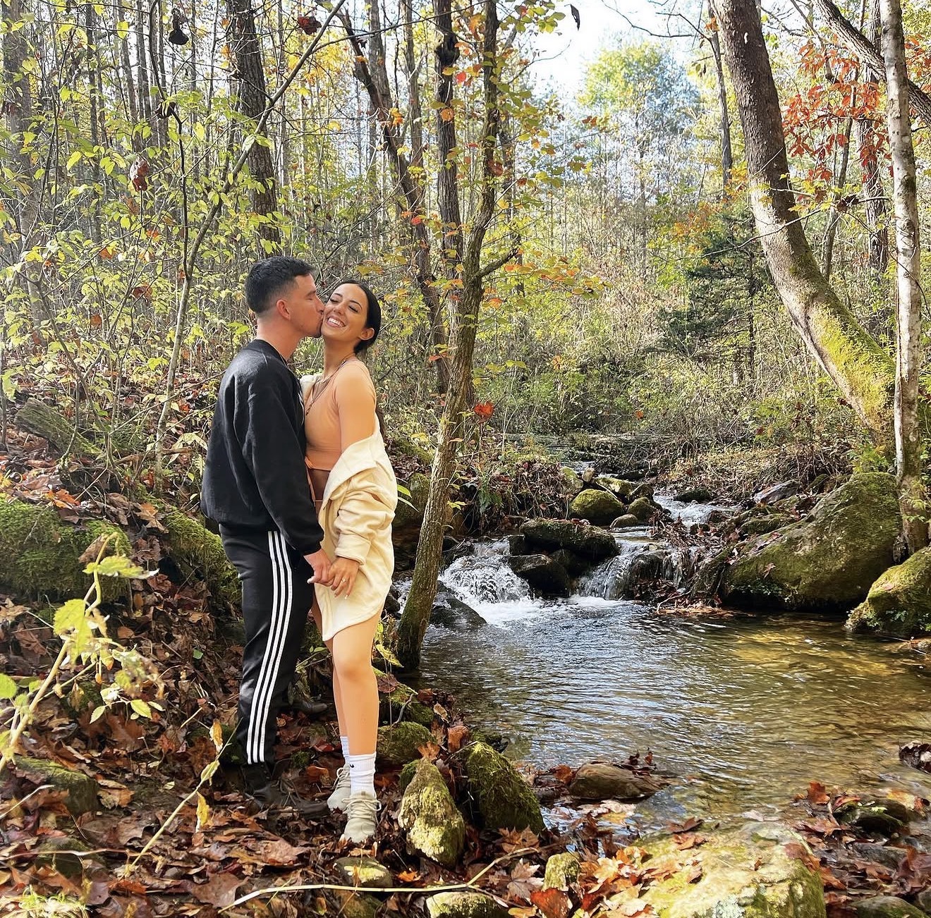 Leon kissing Annalyse on the cheek beside a small waterfall in autumn woods
