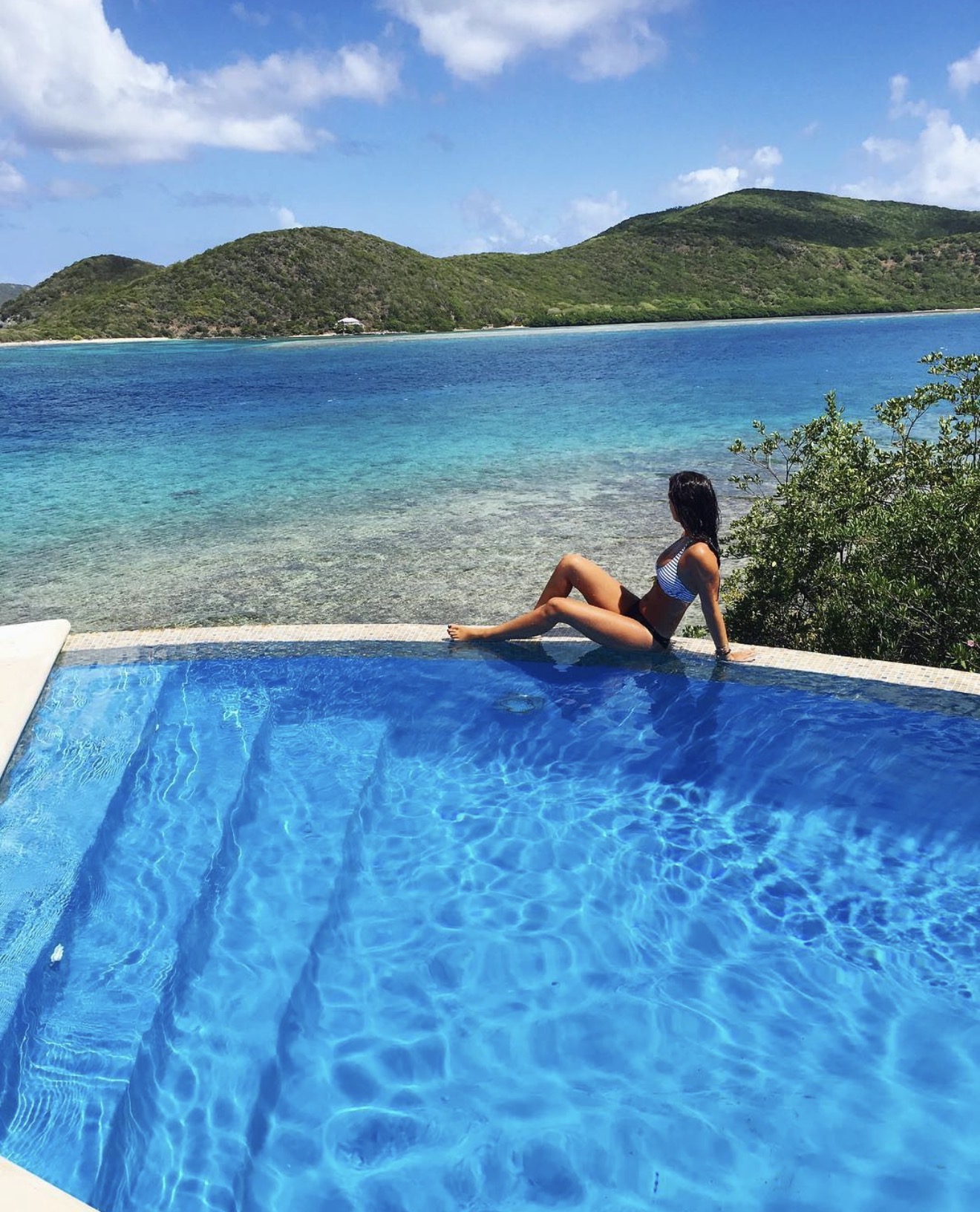 Annalyse sitting on the edge of an infinity pool above turquoise water and green island hills